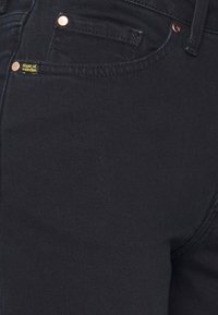 I jeans in denim neri presentano una vestibilità slim, hardware in tonalità rame, una tasca per le monete e un'etichetta gialla sul davanti. Texture liscia, dettagli minimi.