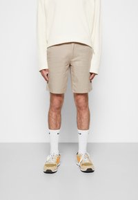 Beige shorts van een lichtgewicht stof, met een lengte tot halverwege de dij. Gecombineerd met een crèmekleurig shirt met lange mouwen, witte sokken en sneakers met oranje accenten.