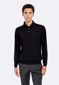 Polo shirt nero a maniche lunghe realizzato in tessuto morbido. Presenta un colletto, una patta con tre bottoni e polsini aderenti. Abbinato a pantaloni grigi.