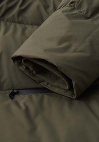 Detail einer olivgrünen Steppjacke mit hochgekrempeltem Bündchen aus weichem Strickmaterial und einer schwarzen Reißverschlusstasche nahe der Naht.