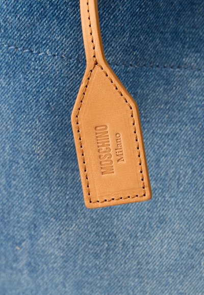 Ετικέτα από ανοιχτό καφέ δέρμα με ανάγλυφο το "MOSCHINO Milano" συνδεδεμένη σε μπλε τζιν ύφασμα με ευδιάκριτη ραφή.