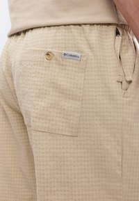 Shorts beiges texturés avec une poche arrière boutonnée et une étiquette de la marque Columbia, portés avec une chemise beige assortie.