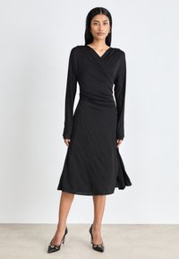Black long-sleeve wrap dress with a V-neck and ruched waist. Soft fabric falls to knee-length, featuring a flowing silhouette and side slits.