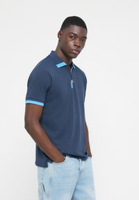 Marineblå polo t-shirt med korte ærmer, prydet med lyseblå accenter ved kraven og ved ærmekanten, lavet af et glat materiale. Lynlåsdetalje ved halsen.