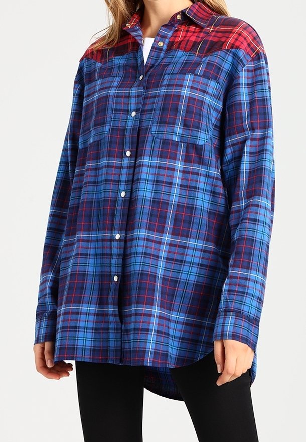 Femme portant une chemise à carreaux bleu et rouge oversize à boutons, manches longues et deux poches poitrine, associée à un pantalon noir.