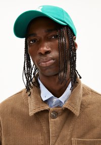 Green cap with embroidered logo, paired with a corduroy brown jacket and a light blue collared shirt. Textured materials highlighted.