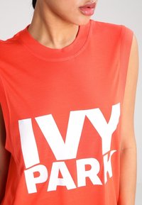 Rød ærmeløs T-shirt lavet af bomuld, med stor hvid tekst "IVY PARK" på forsiden. Glat tekstur med rund halsudskæring.