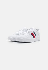 Białe skórzane sneakersy o gładkiej fakturze, z trzema skośnymi paskami w kolorze czerwonym i granatowym. Zapięcie na sznurówki i gumowa podeszwa.