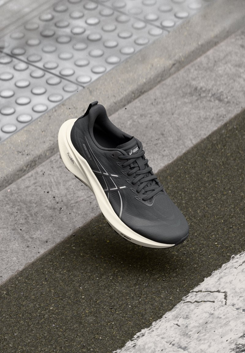 Chaussure de course noire avec semelle blanche flottant au-dessus de l'asphalte près d'un bord en béton et d'un revêtement tactile.