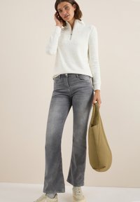 Sweater zippé blanc avec des speckles, jeans gris évasés et baskets beiges. Tenant un sac rembourré vert olive. Fond neutre.