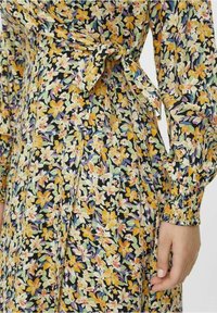 Kwiatowa sukienka z żółtymi, białymi i zielonymi wzorami na czarnym tle. Posiada wiązanie oraz marszczone mankiety na długich rękawach.