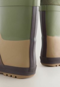 Grüne und beige Gummistiefel mit einer strukturierten Oberfläche, die einen dunkelbraunen Rand und eine gerippte Sohle für besseren Halt aufweisen.