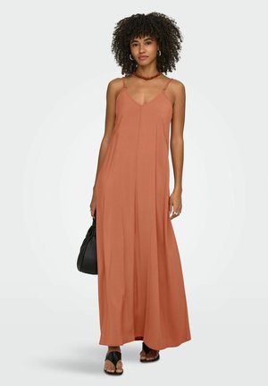 Vrouw met krullend haar die een mouwloze maxi-jurk in roestkleur draagt, zwarte sandalen, een rode ketting, creolen en een zwarte handtas vasthoudt.