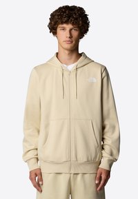 Haut à capuche zippé beige en tissu doux, avec un logo sur le devant, des poches spacieuses et une capuche avec cordon de serrage. Texture lisse, coupe décontractée.