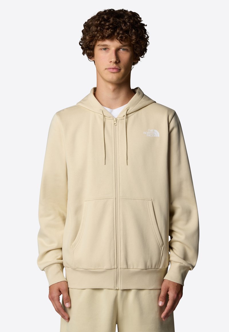 Haut à capuche zippé beige en tissu doux, avec un logo sur le devant, des poches spacieuses et une capuche avec cordon de serrage. Texture lisse, coupe décontractée.