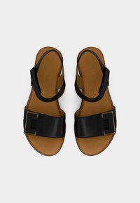 Zwarte leren sandalen met een brede band voorzien van een rechthoekige gouden gesp, een bruine binnenzool en een open teen ontwerp.