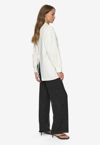 White long-sleeve shirt with a tie at the back, paired with black wide-leg pants featuring white polka dots, and black slip-on sandals.