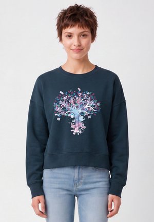 Femme aux cheveux courts portant un sweat-shirt bleu foncé avec un motif d’arbre coloré et un jean bleu clair, debout et faisant face à l’avant.