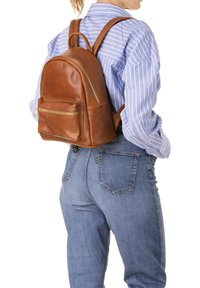 Zaino in pelle marrone con forma arrotondata, tasche con cerniera e spallacci regolabili. Presenta una texture liscia e minimo hardware.