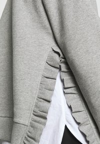 Grauer Sweatshirt mit geripptem Saum und vertikaler Rüschendetails an der Seite, über einem weißen Shirt getragen. Strukturierter Stoff mit glatter Oberfläche.