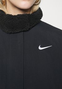 Svart jacka med en fluffig svart krage, med en vit Nike swoosh-logotyp på bröstet. Slät tyg med synliga sömnadsdetaljer.