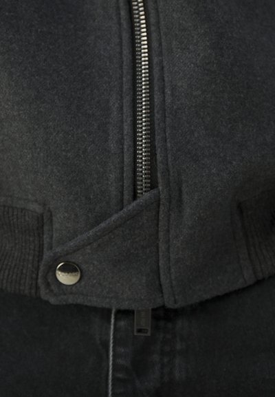 Chaqueta gris oscuro con cierre de cremallera y botón prominentes, con acentos de canalé. El material parece suave y texturizado.