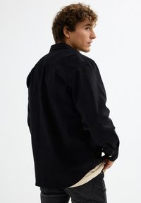 Camisa negra de botones en un ajuste relajado, con cuello, mangas largas y una textura suave, combinada con una camiseta interior clara.