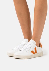 Baskets blanches avec des accents orange, fabriquées en cuir lisse, dotées d'une pointe arrondie, d'un laçage plat et d'une semelle en caoutchouc texturé.