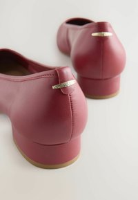 Bailarinas de imitación cuero rojas con punta redondeada, tacón bajo y acento metálico en la parte trasera. Textura suave y detalles de costura mínimos.