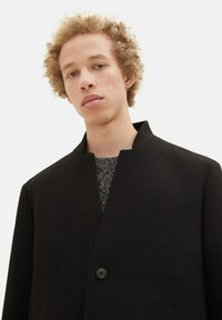 Schwarzer Mantel mit Stehkragen, einem Knopf und glatter Textur, kombiniert mit einem grauen strukturierten Pullover. Das Model hat lockiges Haar.