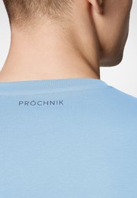 Jasnoniebieska koszulka z miękkiego materiału, z okrągłym dekoltem. Na plecach znajduje się nadruk w ciemnoszarym kolorze z logo marki "PROČHNIK".