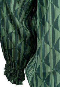 Manche de blouse verte transparente avec motif géométrique circulaire et rayé, poignet smocké élastique créant un détail froncé.