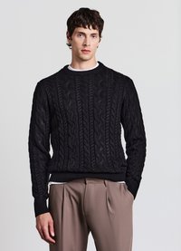 Schwarzer Strickpullover mit Zopfmuster, rundem Ausschnitt und gerippten Bündchen; getragen über einem weißen Shirt mit Rundhalsausschnitt, kombiniert mit beigen Hosen.