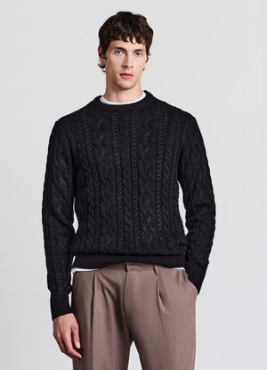 Schwarzer Strickpullover mit Zopfmuster, rundem Ausschnitt und gerippten Bündchen; getragen über einem weißen Shirt mit Rundhalsausschnitt, kombiniert mit beigen Hosen.
