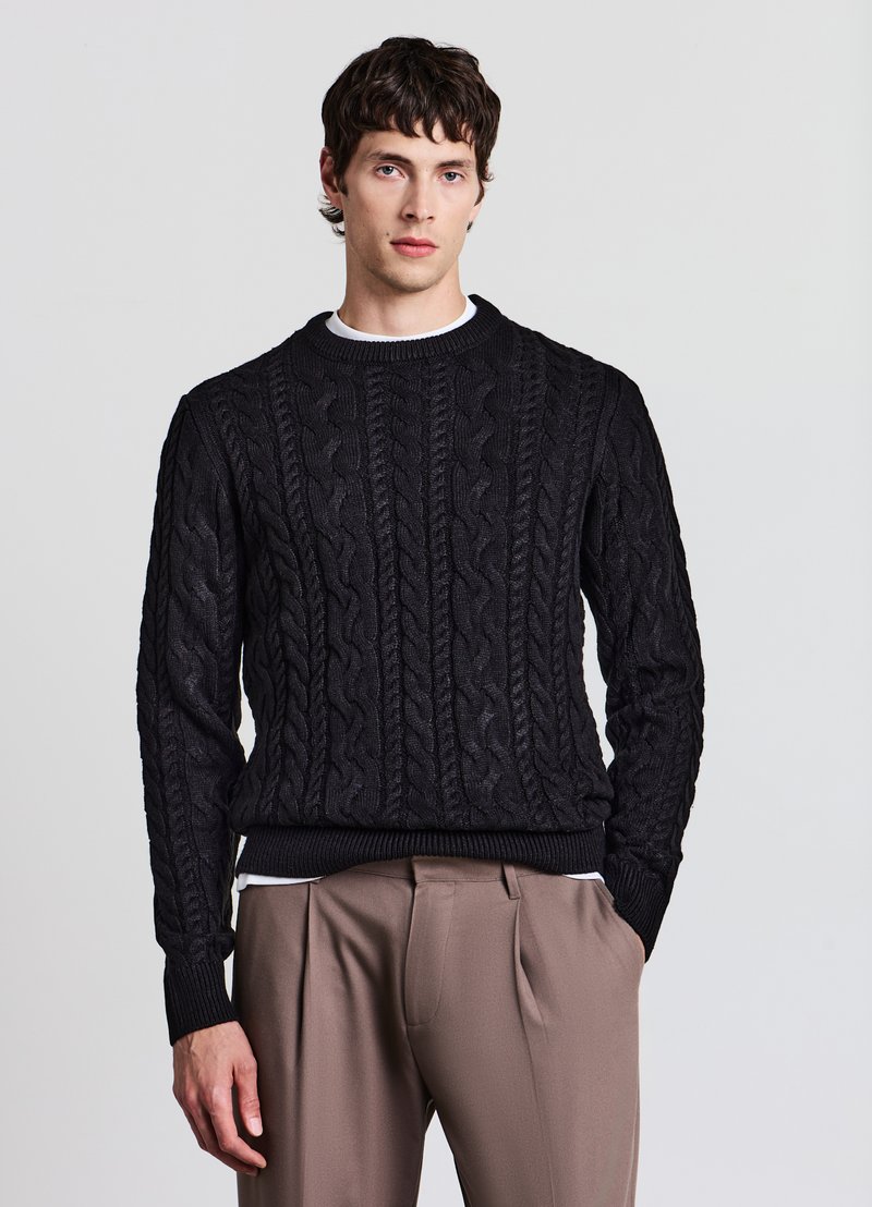 Schwarzer Strickpullover mit Zopfmuster, rundem Ausschnitt und gerippten Bündchen; getragen über einem weißen Shirt mit Rundhalsausschnitt, kombiniert mit beigen Hosen.