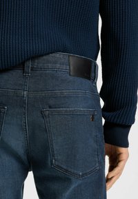 Dark blue denim jeans with a smooth texture, featuring a black leather waistband patch and two back pockets. Knitted navy sweater visible above.