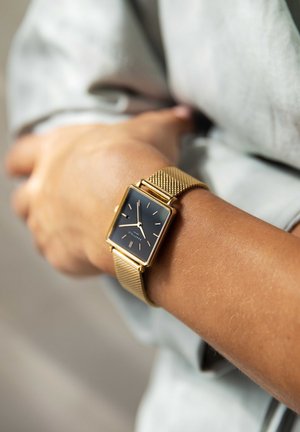 Reloj con correa de malla dorada y una esfera cuadrada negra, que presenta marcadores horarios minimalistas y un diseño elegante. Lleva puesto en la muñeca de una persona.