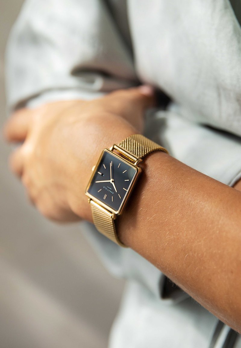 Gouden mesh strap horloge met een vierkante zwarte wijzerplaat, met minimalistische uurmarkers en een elegant ontwerp. Draagt op de pols van een persoon.