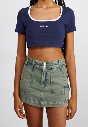 Femme portant un top court Nike bleu marine avec un bord blanc et une mini-jupe en jean vert olive avec des poches avant et une fermeture à double bouton.