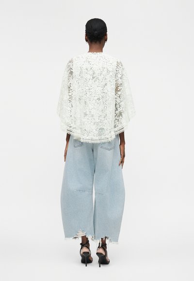 Femme debout, dos tourné, portant un top cape en dentelle florale blanche, un jean large déchiré bleu clair et des sandales noires à talons.