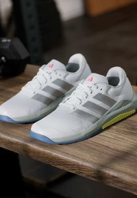Białe sneakersy Adidas z szarymi paskami, różowym logo oraz niebiesko-żółtymi podeszwami, ułożone obok siebie na drewnianej powierzchni.