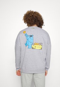 Sweatshirt gris en coton, comportant un personnage de cartoon bleu et un graphique de basket-ball dans le dos, avec des poignets et un ourlet côtelés.