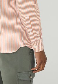 Chemise rayée orange et blanche avec des poignets boutonnés, associée à un pantalon cargo vert olive avec un détail de poche boutonnée.