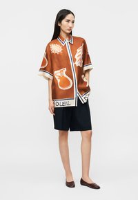 Femeie purtând o cămașă maro largă cu imprimeuri de soare și ulcior, pantaloni scurți bleumarin și pantofi maro, stând în picioare pe un fundal simplu.