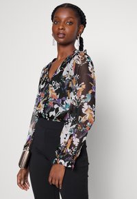 Blusa preta transparente com padrões florais coloridos, mangas longas bufantes, decote em V e punhos ligeiramente franzidos, combinada com calças de cintura alta.