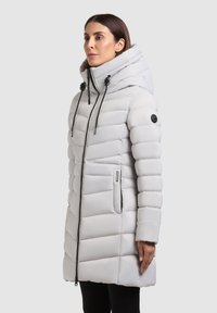 Femeie îmbrăcată cu o haină de tip puffer lungă, matlasată, de culoare gri deschis, cu guler înalt, glugă ajustabilă, închidere cu fermoar și buzunare laterale.