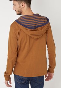 Camisa de manga larga marrón con capucha a rayas azules. Presenta un corte relajado y una tela suave. La capucha tiene un color y un diseño en contraste.