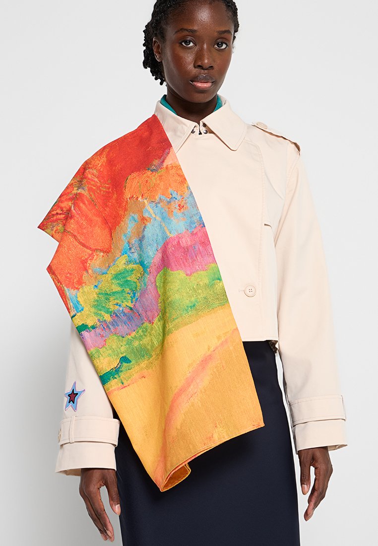 Kobieta w beżowej, krótnej kurtce z dużym, kolorowym abstrakcyjnym szalem przewieszonym przez lewe ramię oraz granatowej spódnicy, stojąca na tle białym.