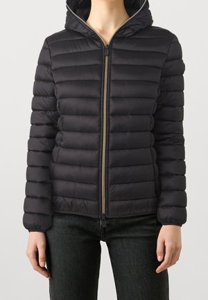 Veste noire matelassée à capuche avec fermeture éclair à l'avant portée par une personne en pantalon noir sur un fond uni.
