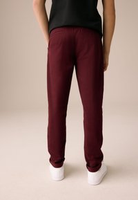 Pantalon fuselé bordeaux en tissu lisse, avec poches arrière et coupe slim, porté avec des baskets blanches.
